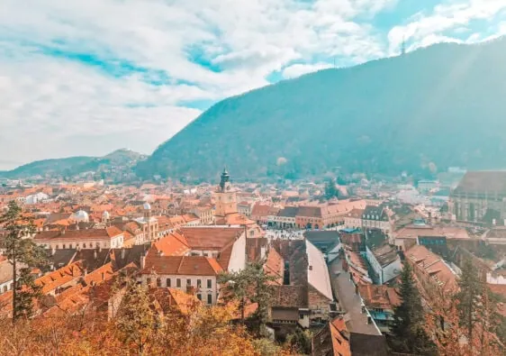 A panoramic view of a European town with terracotta rooftops and pastel-colored buildings, nestled in a valley surrounded by green hills. The town square is visible in the distance, with a clock tower standing prominently in the center under a bright blue sky.