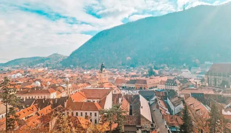 A panoramic view of a European town with terracotta rooftops and pastel-colored buildings, nestled in a valley surrounded by green hills. The town square is visible in the distance, with a clock tower standing prominently in the center under a bright blue sky.