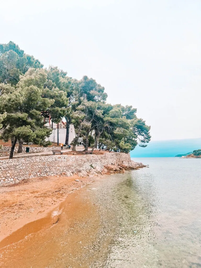 A peaceful seaside scene featuring a narrow sandy beach leading to clear shallow waters. A stone wall lines the edge of the shore, with pine trees and a few people walking along the pathway under an overcast sky.