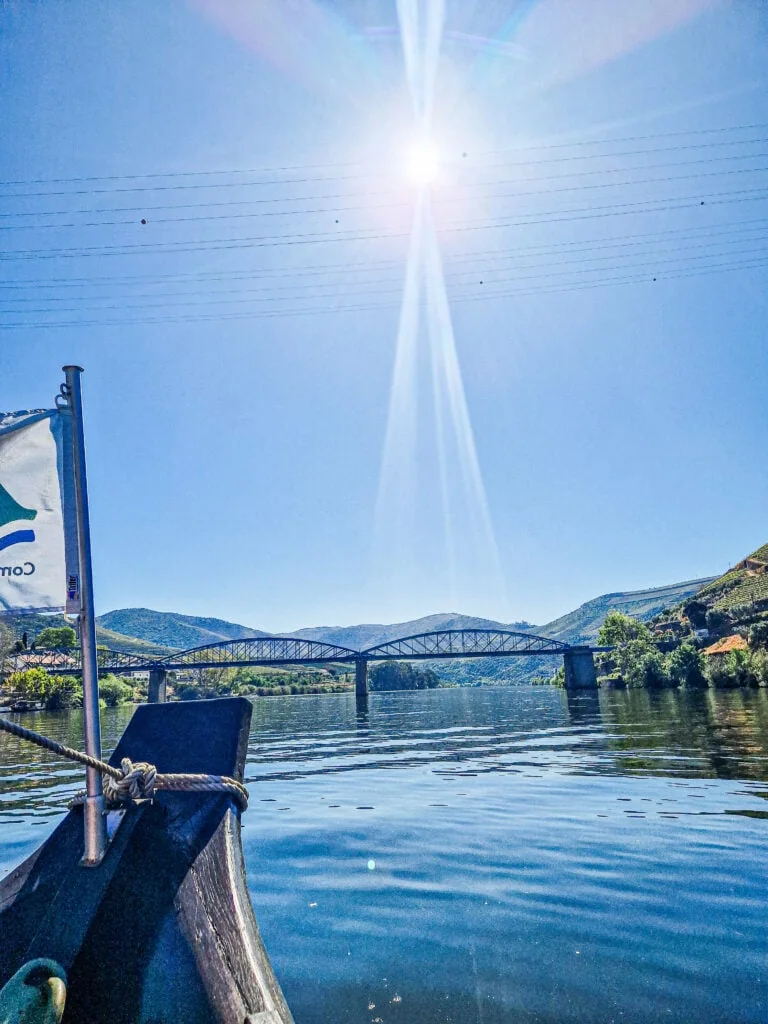 A boat ride along the Douro River, with the tip of a traditional wooden vessel visible in the foreground. A flag with a green and blue logo flutters in the wind, while the sun shines brightly overhead, casting a radiant glow on the water and surrounding hills.