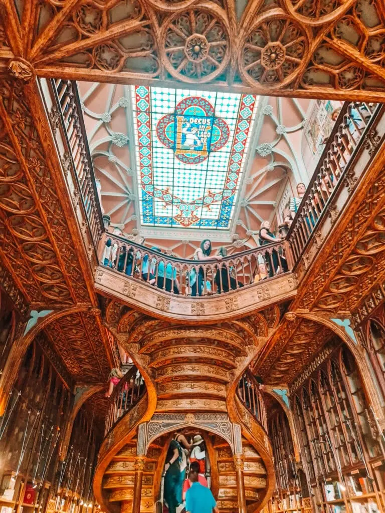 A grand, intricately carved wooden staircase inside a historic bookstore, with visitors admiring the details. A stained-glass ceiling with the Latin phrase "Decus in Labore" adds to the architectural beauty.