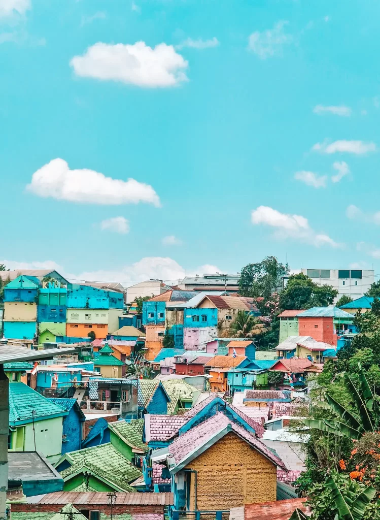 A vibrant hillside neighborhood with houses painted in bright shades of blue, green, yellow, orange, and pink under a clear blue sky with fluffy white clouds. The colorful rooftops and facades create a striking contrast against the greenery and modern buildings in the background.