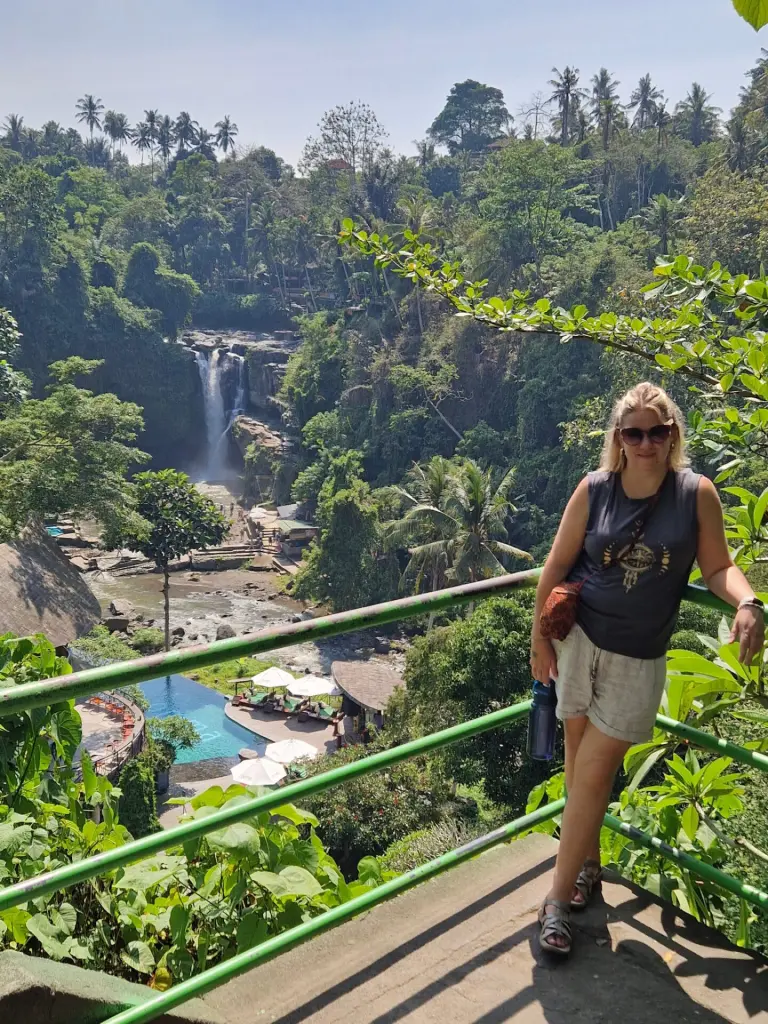 Em wearing sunglasses, a dark tank top, and light-colored shorts leans against a green railing with a waterfall cascading in the background. The lush tropical landscape is filled with dense greenery, palm trees, and a resort-style pool with lounge chairs below. The bright sunlight highlights the scenic beauty, creating a perfect vacation setting.