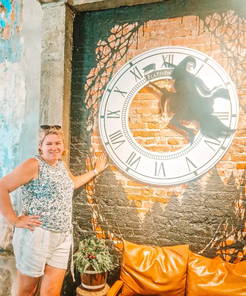 Em poses in front of a large Harry Potter-themed clock mural featuring a witch silhouette on a broomstick, located inside Platform 9 1/2 Café—perfect for fans adding magic to their 1 day in Ipoh adventure.
