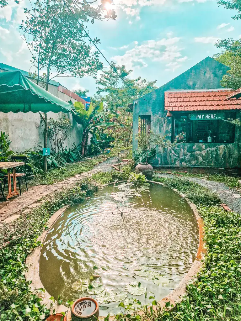A tranquil garden café with a circular lily pond and gentle fountain in the center, surrounded by lush greenery and rustic stone paths. In the background, there's a small café hut with a sign that reads “PAY HERE,” set against a sunny sky with scattered clouds.