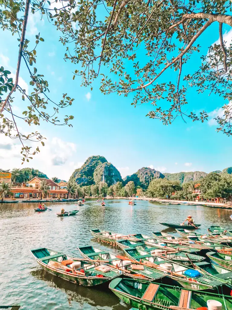 Dozens of green rowboats are docked along the peaceful Tam Cốc river, framed by leafy branches overhead. The backdrop features limestone karst mountains, traditional buildings, and tourists paddling on the water under a bright blue sky.