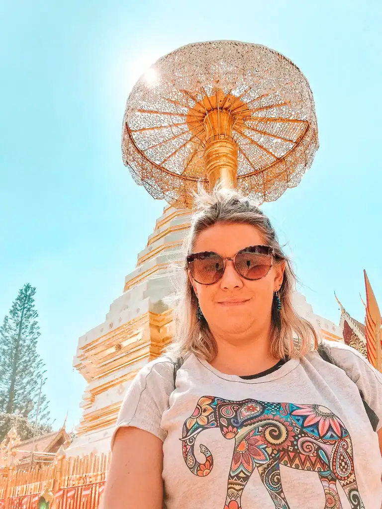 Em wearing sunglasses and a t-shirt with a colorful elephant design smiles in front of the intricate golden umbrella at Wat Phra That Doi Suthep, with bright sunlight streaming through.