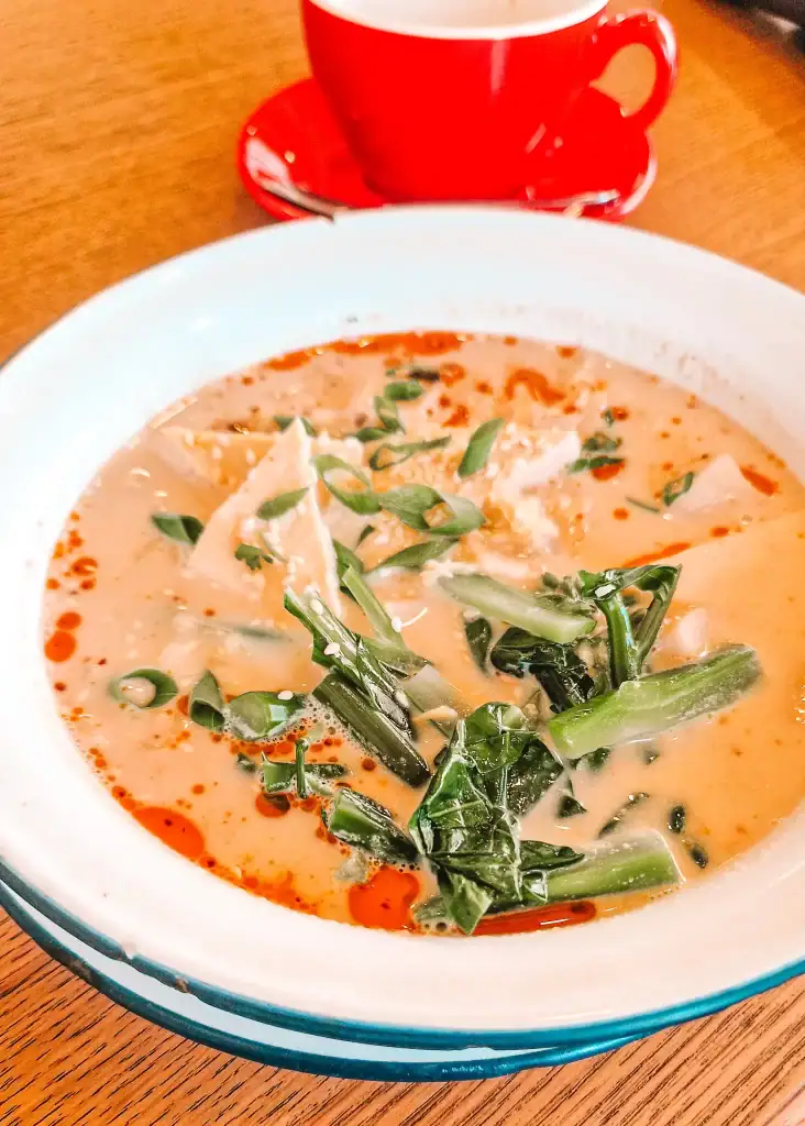 A steaming bowl of miso ramen soup topped with leafy greens, scallions, and dumplings, with a drizzle of chili oil on the creamy broth, served alongside a red cup of coffee on a wooden table.