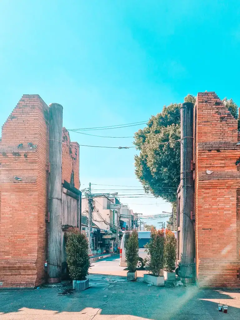 The historic Tha Phae Gate in Chiang Mai, made of red brick and partially crumbling, opens into a busy street lined with buildings, greenery, and power lines under a sunny sky.