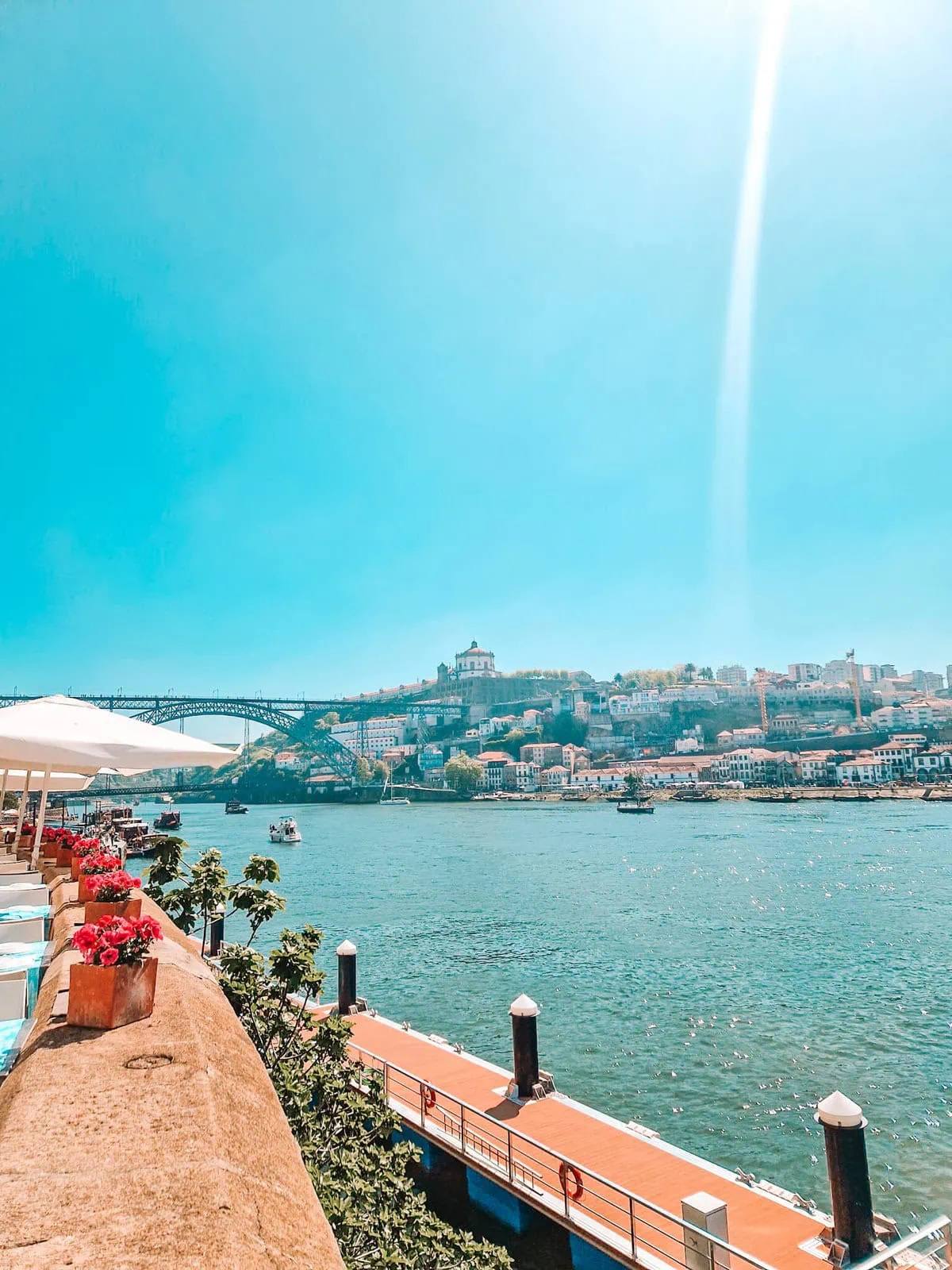 A scenic view of the Douro River in Porto, Portugal, with a riverside café adorned with red flowers. Boats float on the shimmering water, and the Dom Luís I Bridge and historic hillside buildings are visible in the background.