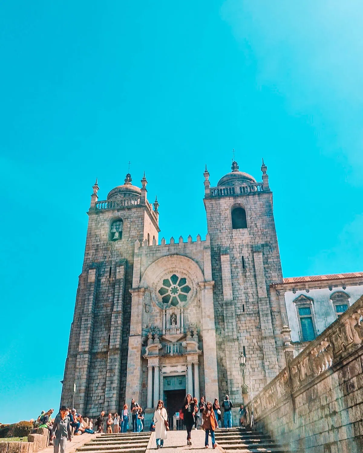  grand, stone-built cathedral with two prominent towers, intricate rose windows, and arched doorways. A bright blue sky serves as the backdrop, while a diverse group of visitors walks up the stone steps, capturing photos and admiring the architecture.
