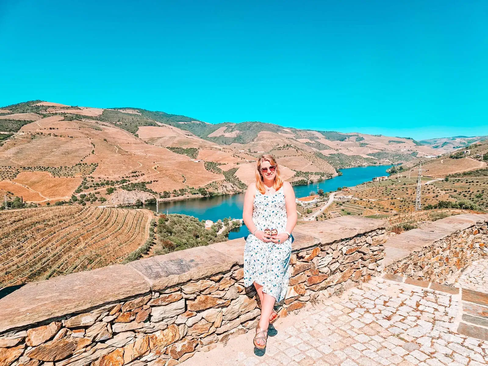 A woman with sunglasses and a light blue floral dress sits on a stone wall overlooking the Douro Valley. The golden vineyards stretch across the hills behind her, with the blue river cutting through the landscape