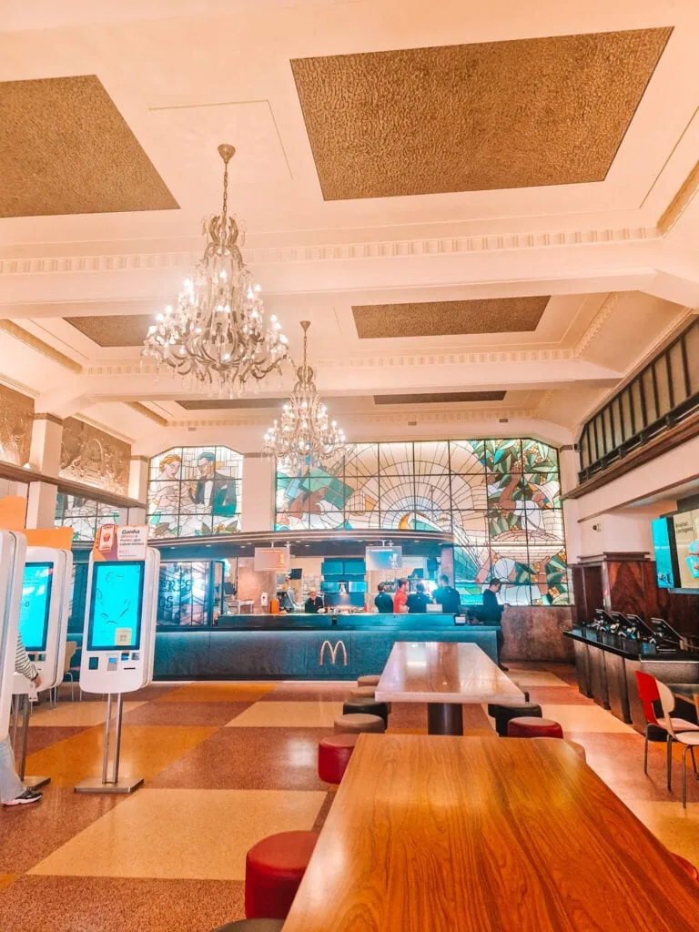 A McDonald's restaurant inside a historic building with ornate chandeliers, stained glass windows, and high ceilings. Wooden tables, modern ordering kiosks, and a counter with employees serving customers blend fast food with a luxurious setting.