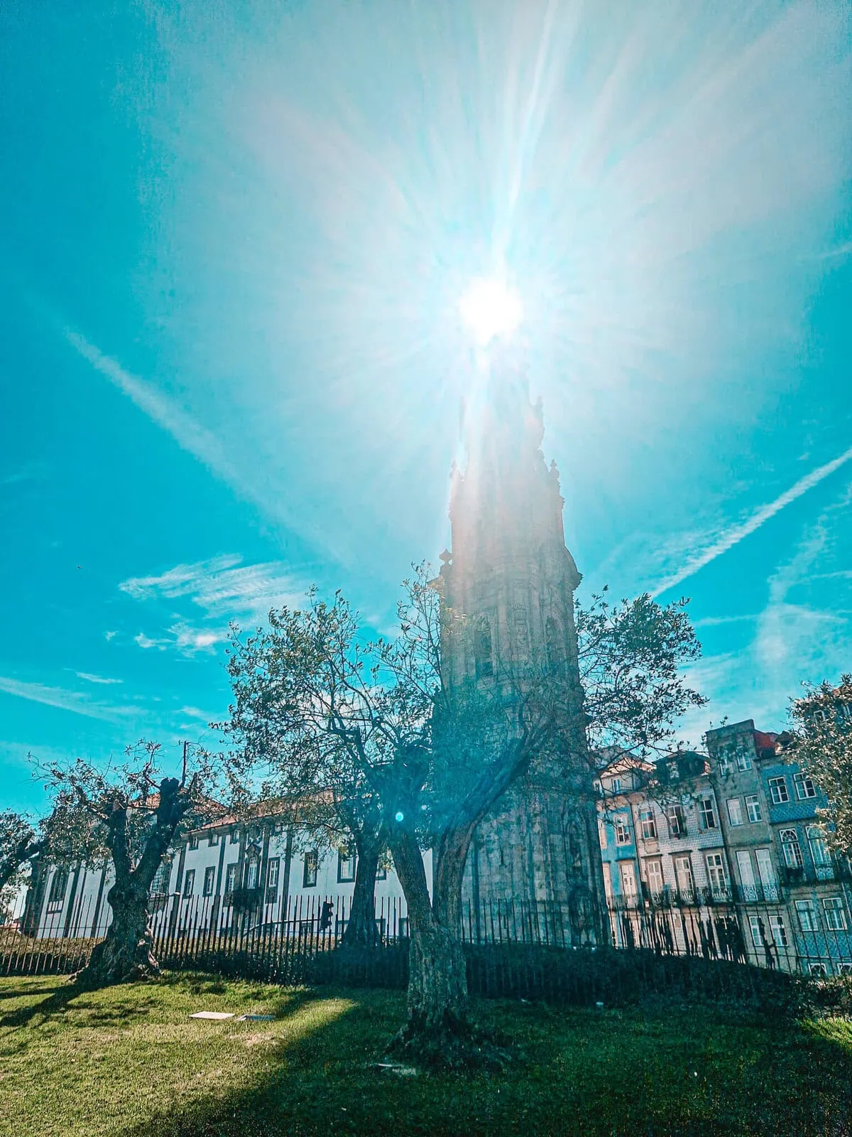 A historic stone tower stands tall in a green park, with old, twisted trees surrounding it. The bright sun shines directly behind the tower, creating a dramatic lens flare effect against the vivid blue sky.