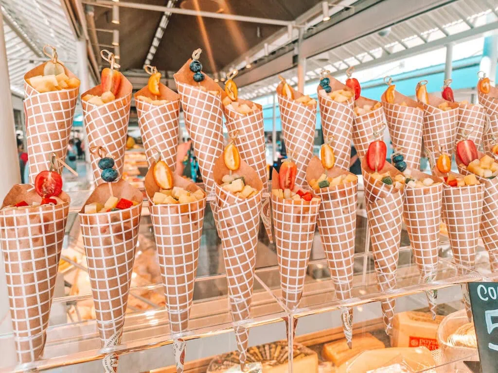 A display of waffle cones filled with fresh fruit, cheese, and cherry tomatoes, each cone wrapped in a checkered paper and topped with a skewer of blueberries, grapes, or tomatoes.