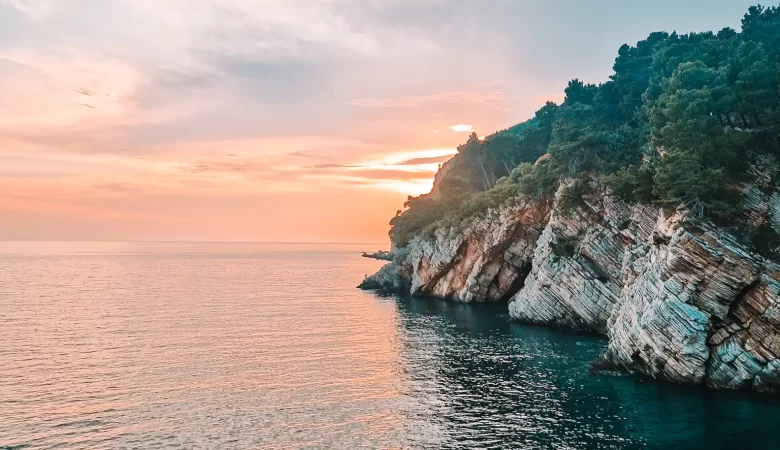 A peaceful seascape at sunset, with the sky painted in soft pastel hues. The calm water meets rugged cliffs covered in lush green trees, as the last rays of sunlight peek over the horizon.