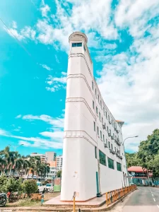 A uniquely shaped, narrow white building with a curved tower-like corner stands against a bright blue sky with scattered clouds. The surrounding area includes palm trees, modern buildings, and a street with parked cars.