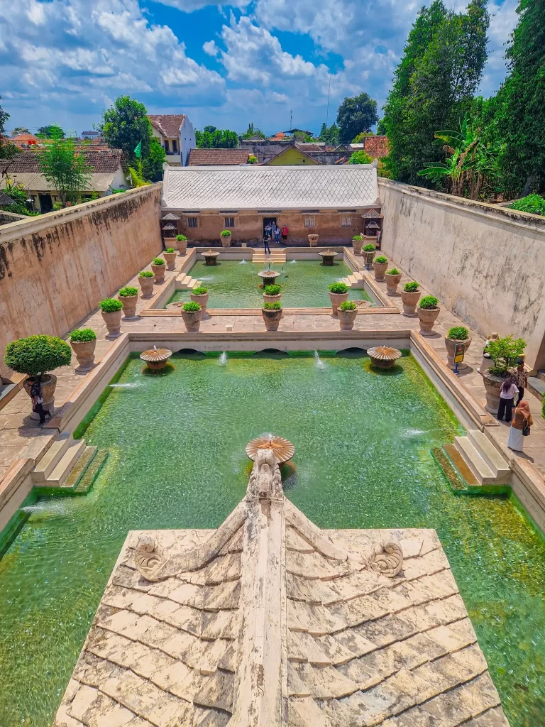 Overhead view of two large green pools separated by a bridge, surrounded by walls with potted plants