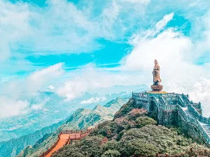 A majestic bronze statue stands on a mountaintop platform, surrounded by clouds. A long red pathway and staircases lead up to the monument, offering breathtaking views of the mountain range below.