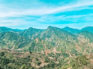 A panoramic landscape of green mountains with winding dirt roads and terraced rice fields stretching across the slopes. The sky is bright blue with wispy clouds, and the valleys below are covered in dense vegetation.