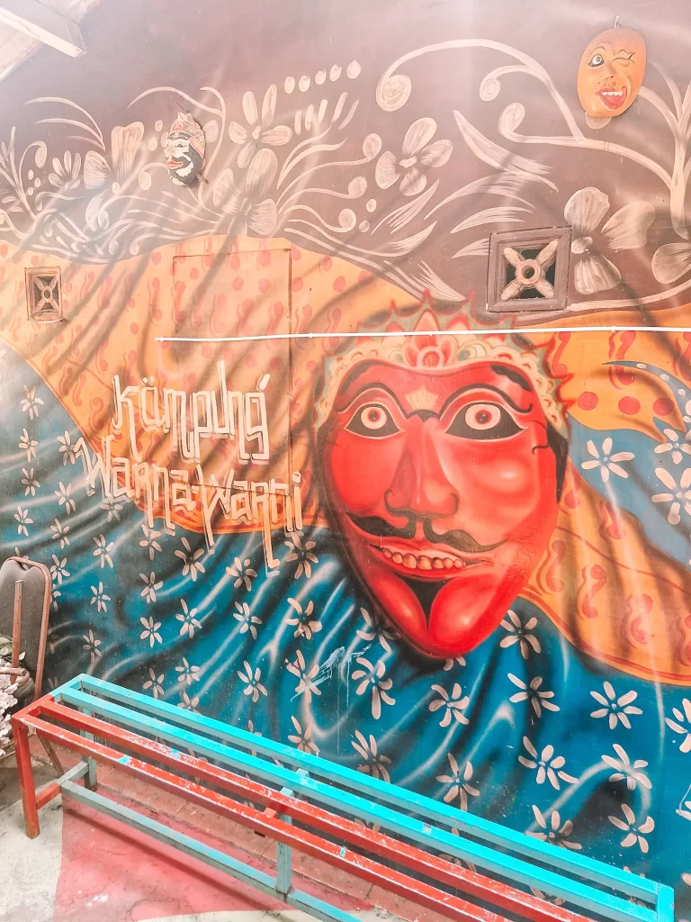 A vibrant mural featuring a large red mask with exaggerated facial features, set against a patterned blue and gold background. The words "Kampung Hanavarti" are painted in bold letters, blending into the intricate design. A wooden bench with turquoise and red slats sits in front of the artwork.