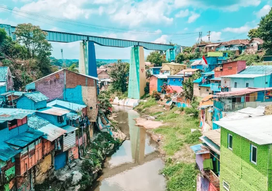 A vibrant riverside neighborhood with houses painted in bright blue, green, and other bold colors, lining the edge of a murky river. A large elevated bridge with green pillars stretches across the background, connecting two areas. The sky is bright blue with fluffy white clouds, adding to the colorful and lively atmosphere of the scene.