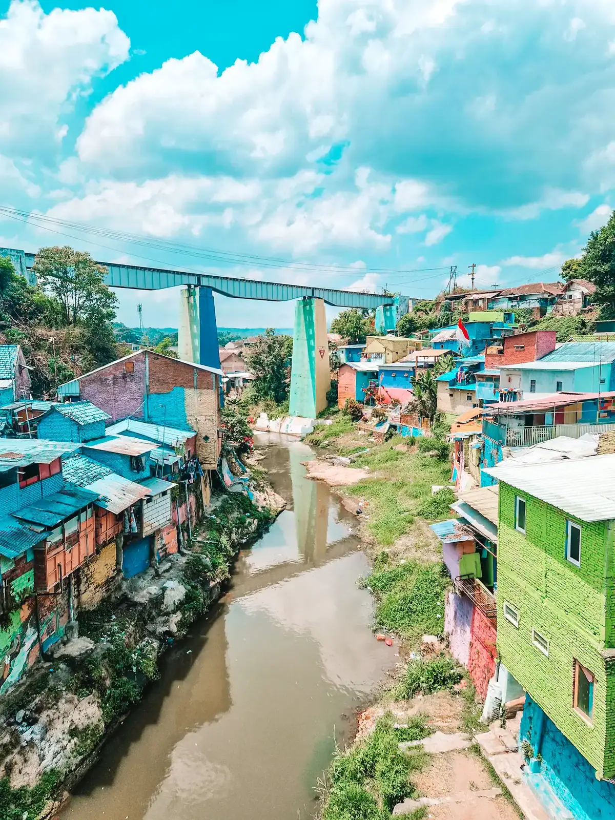 A vibrant riverside neighborhood with houses painted in bright blue, green, and other bold colors, lining the edge of a murky river. A large elevated bridge with green pillars stretches across the background, connecting two areas. The sky is bright blue with fluffy white clouds, adding to the colorful and lively atmosphere of the scene.