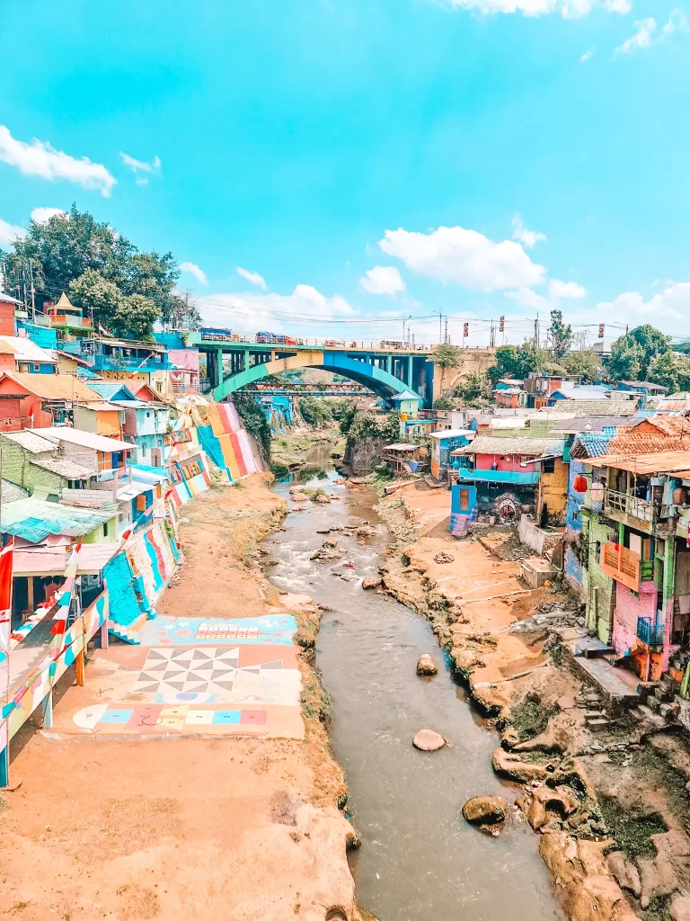 A vibrant riverside community with houses painted in bold colors, including blues, yellows, and reds, sitting along the banks of a shallow, rocky river. A large, multicolored bridge spans the background, while painted murals and geometric patterns decorate the riverbanks, adding to the lively atmosphere under a bright blue sky.