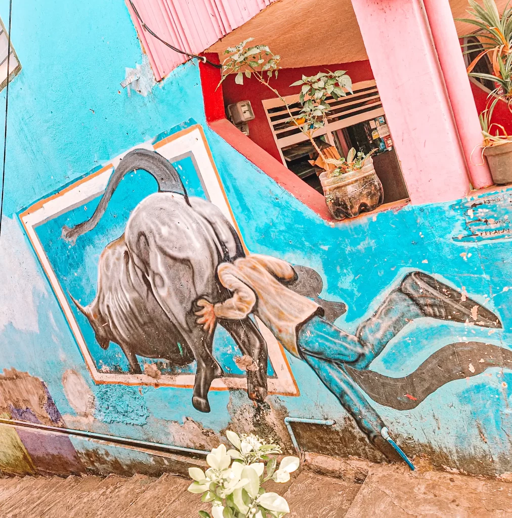 A 3D-style street mural showing a man appearing to be pushed out of a picture frame by a charging bull. The illusion makes it look as if the bull is bursting out of the wall, knocking the man backward. The surrounding area has bright blue walls, pink accents, and potted plants adding to the lively scene.