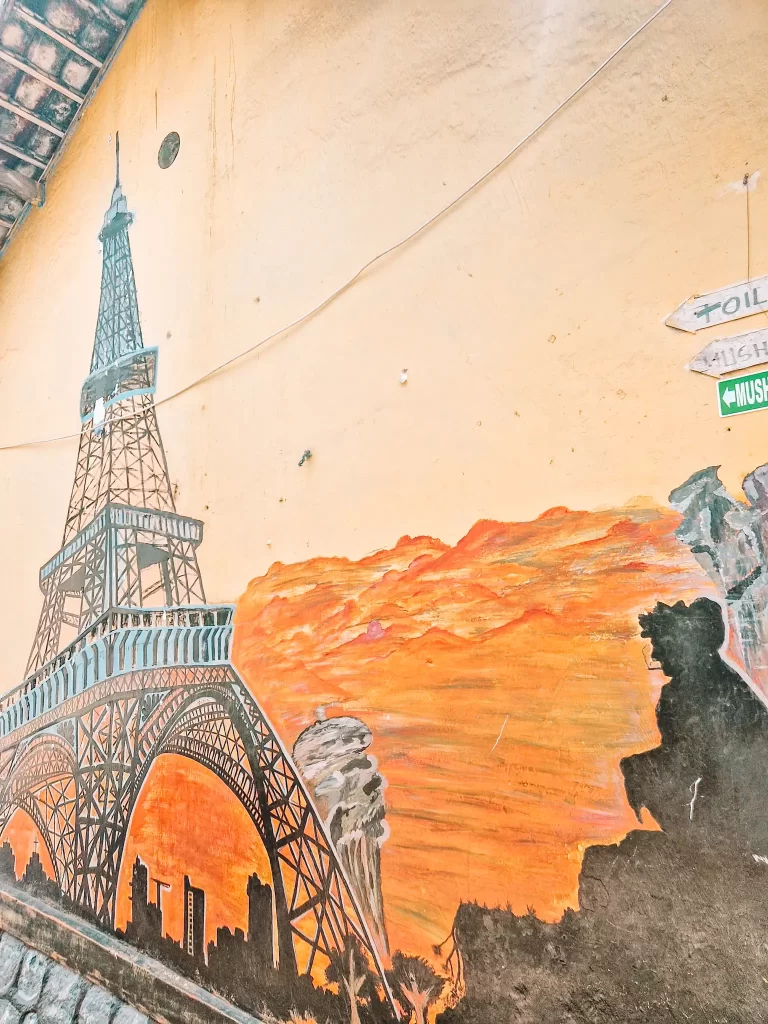 A detailed mural of the Eiffel Tower set against a striking orange sunset. The lower part of the mural shows a city skyline in silhouette, while the warm colors blend with the texture of the wall, creating a rustic and artistic effect.