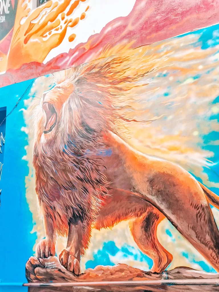 A vibrant mural of a roaring lion standing on a rocky surface, with its mane flowing dramatically in the wind. The background features a bright blue sky and dynamic splashes of orange and red, adding to the powerful energy of the artwork.