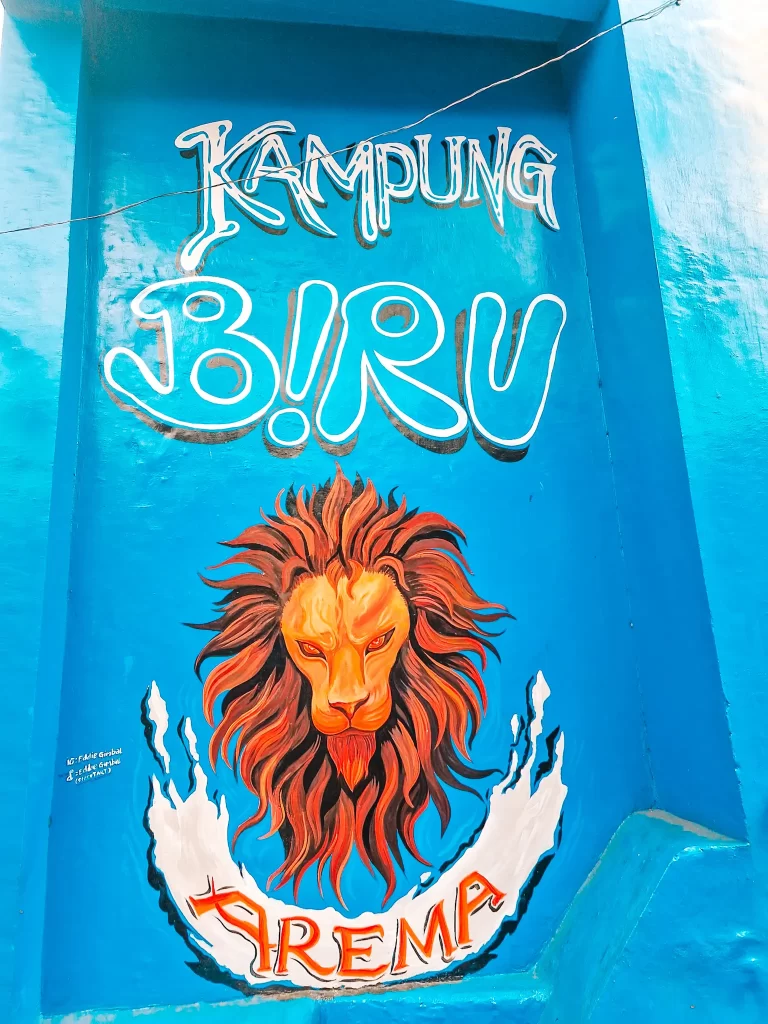 A bright blue mural with the words "Kampung BIRU Arema" features a detailed painting of a lion's head with a fiery mane, surrounded by white, wave-like shapes. The bold graffiti-style lettering and vivid colors make the mural stand out.