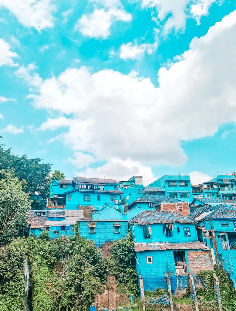 A hillside neighborhood with houses painted in various shades of blue, creating a striking contrast against the lush green vegetation and bright blue sky with fluffy white clouds. Some homes have tiled roofs, while others have exposed brick elements, adding to the rustic charm.