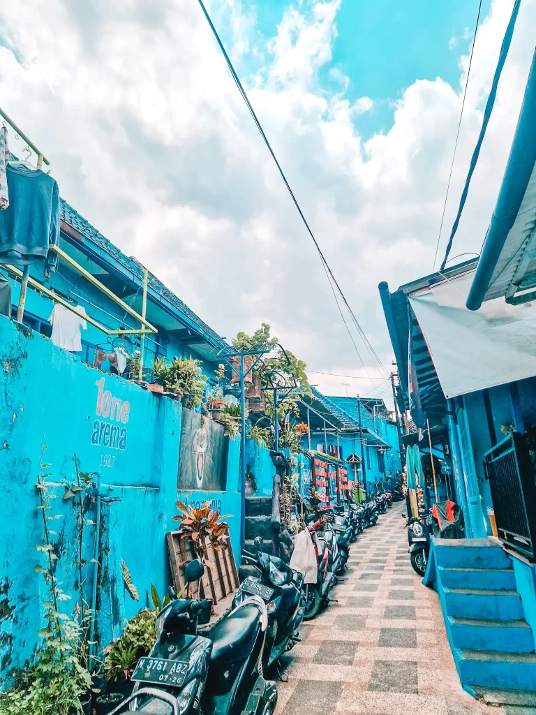 A vibrant alleyway in a neighborhood painted entirely in blue, known as "Tone Arema 1987." Rows of motorbikes are parked along the checkered pavement, while potted plants and colorful murals add character to the space. The sky above is bright with scattered clouds.