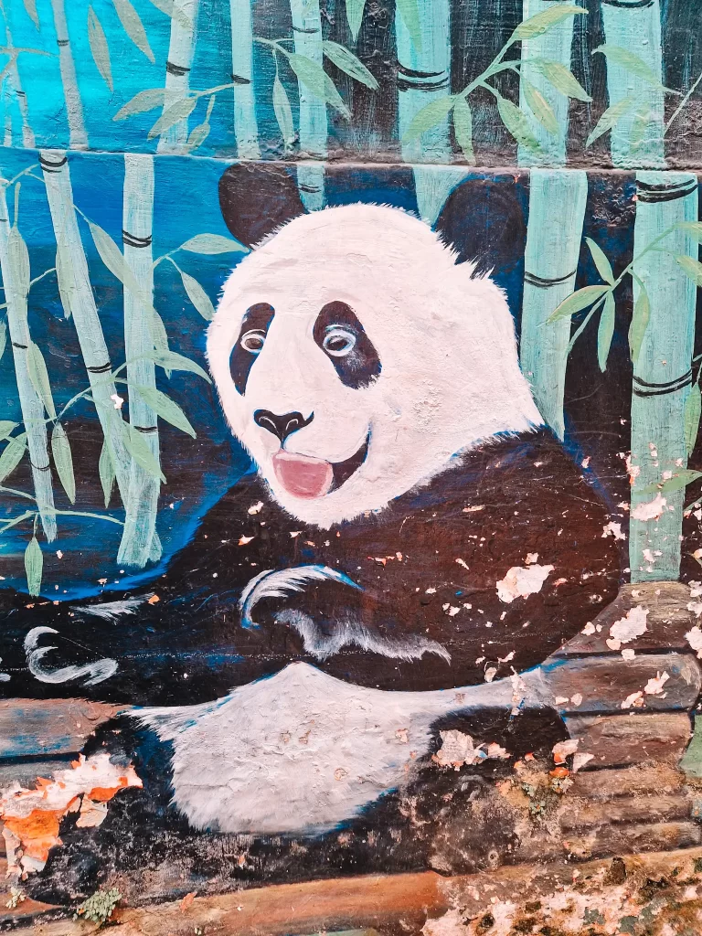 A colorful mural of a panda surrounded by bamboo, painted on a weathered wall. The panda has a playful expression with its mouth open, and some of the paint is chipped, revealing the underlying surface. The green bamboo stalks contrast with the bright blue background.