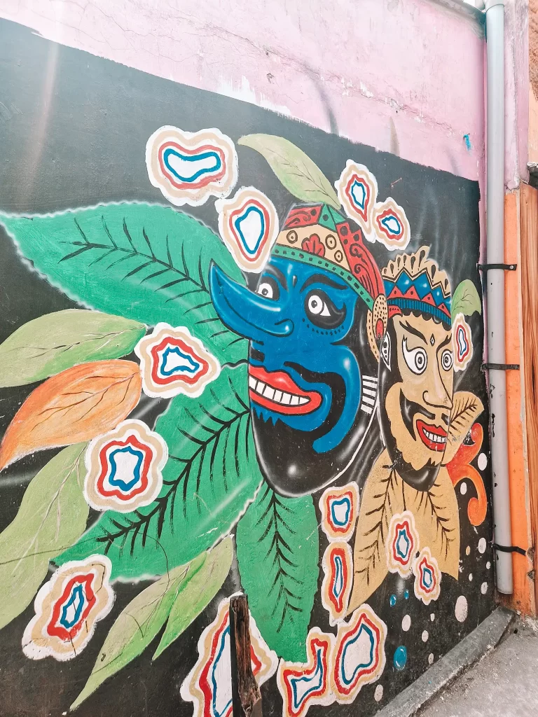 A colorful street mural featuring two traditional masks with exaggerated features—one blue with a long nose and sharp teeth, and the other beige with a wide, toothy grin. The masks are surrounded by green leaves and abstract, layered patterns in white, orange, and blue, giving the mural a lively and cultural aesthetic.