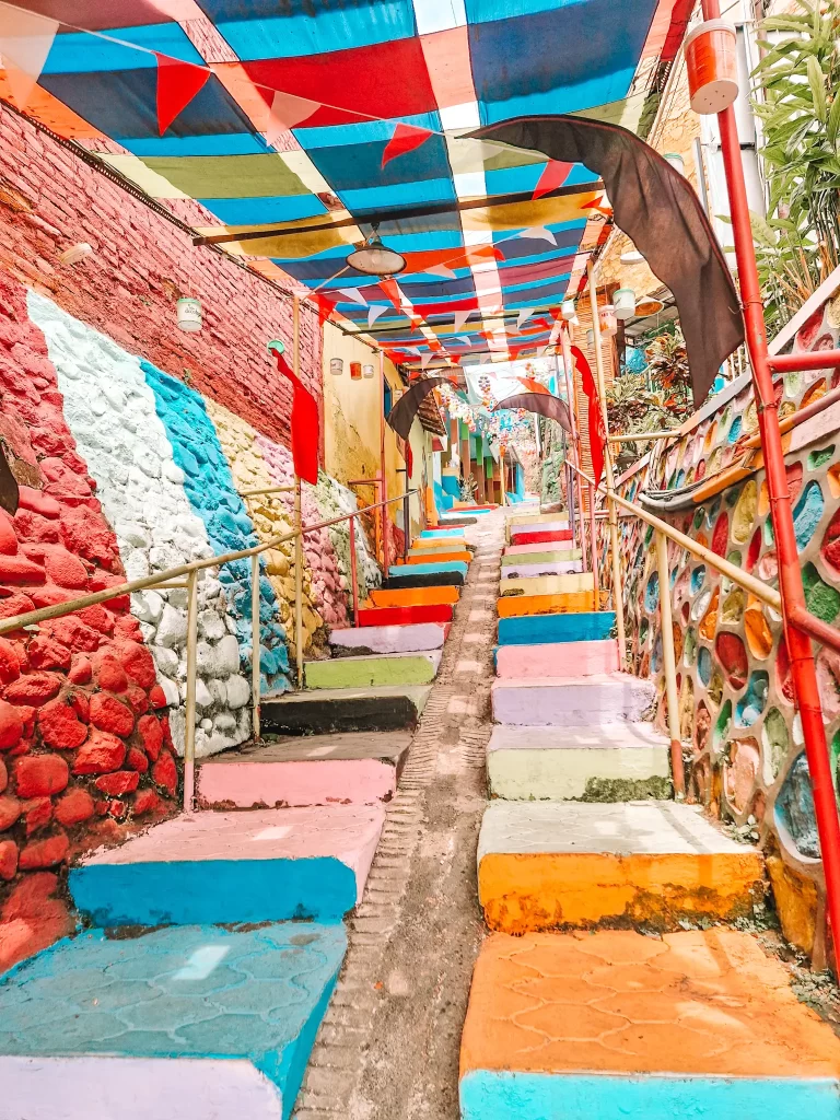 A staircase painted in bright, multicolored steps ascends through a narrow alleyway, covered by a canopy of patchwork blue, red, and yellow fabric. The walls on either side are adorned with textured, colorful murals, creating a lively and artistic atmosphere.