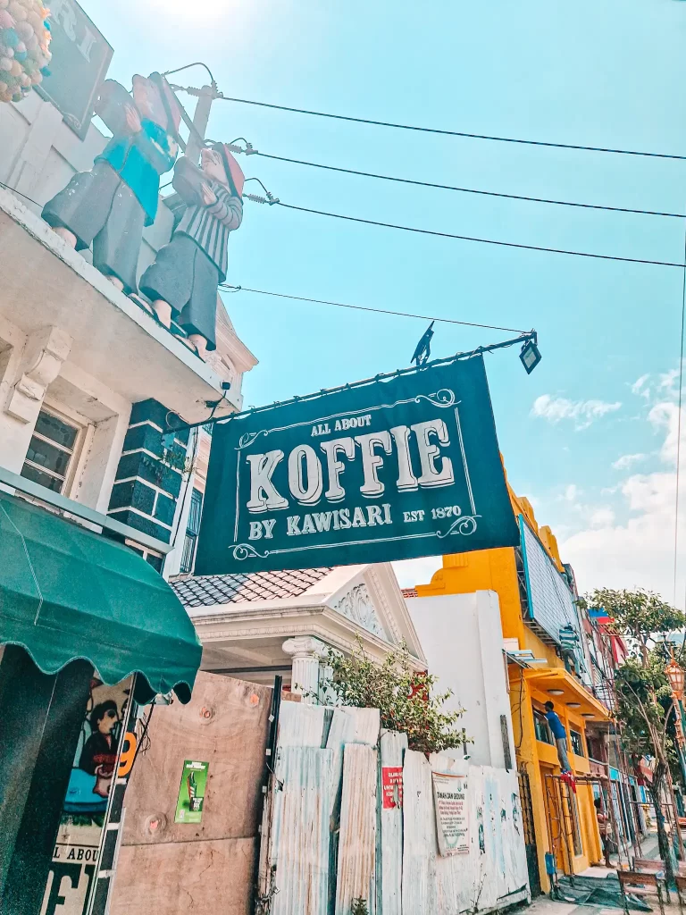 A café sign reading "All About Koffie by Kawisari Est. 1870" hangs over a lively street. The building has a European-style façade, with two statues of people in traditional clothing positioned on the rooftop. The street below is colorful, with bright yellow and blue-painted walls.