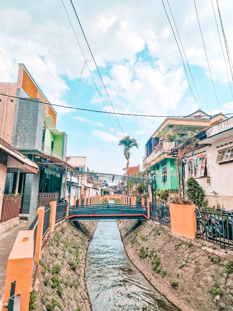 A narrow canal runs through a residential area, lined with houses painted in shades of green, white, and beige. A small pedestrian bridge with red and black railings spans the canal, while overhead power lines crisscross the bright blue sky.