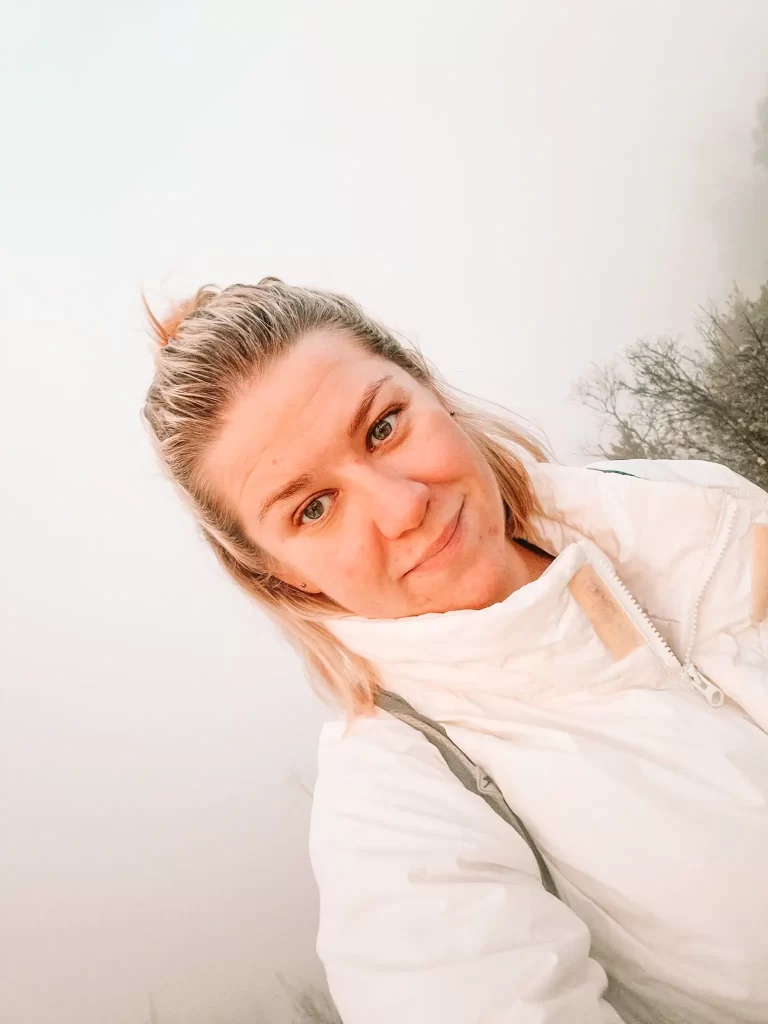 Em in a white puffer jacket takes a selfie in a foggy outdoor setting. She has a soft smile and slightly windblown hair. The misty background gives the scene an ethereal atmosphere, with faint outlines of trees visible behind her.