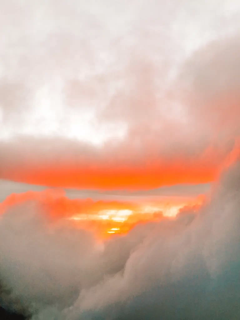 A stunning sunrise emerges through thick clouds, casting a warm orange and red glow over the sky. The sun’s rays filter through the mist, creating a dramatic contrast between the dark clouds and the fiery hues of the morning light. The view is partially obscured by the surrounding fog.