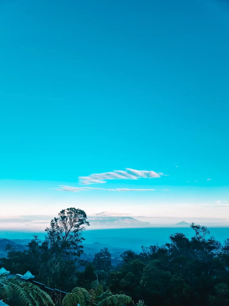 A breathtaking view of a mountain range at sunrise, with a clear blue sky and wispy clouds above. Layers of mist settle over the distant peaks, creating a serene and dreamy effect. Tall trees in the foreground frame the scene, adding depth to the landscape.