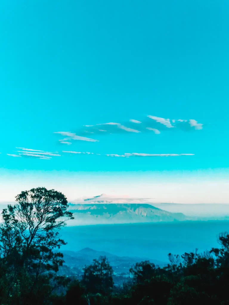 A stunning view of the Mount Bromo landscape with a clear blue sky, distant mountain peaks, and lush greenery—an essential stop in any Mount Bromo itinerary.