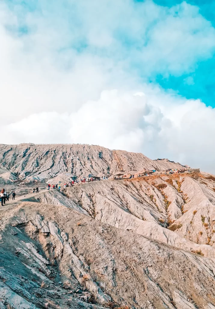 A rugged volcanic landscape with a steep trail leading up to the crater. A line of hikers and visitors makes their way up the rocky path, with fluffy clouds and a vibrant blue sky above.