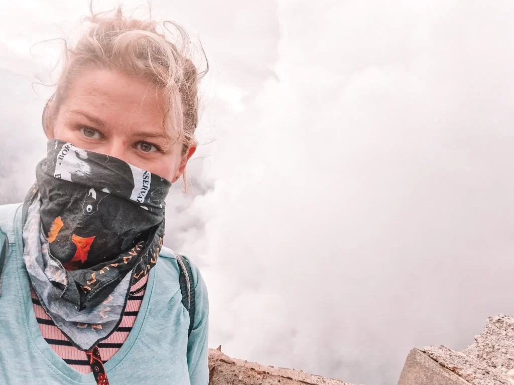 A woman wearing a blue shirt and a patterned bandana covering her mouth, standing near the edge of a volcanic crater with thick white smoke rising behind her. Her eyes are focused on the camera, and the background is hazy from the volcanic activity.