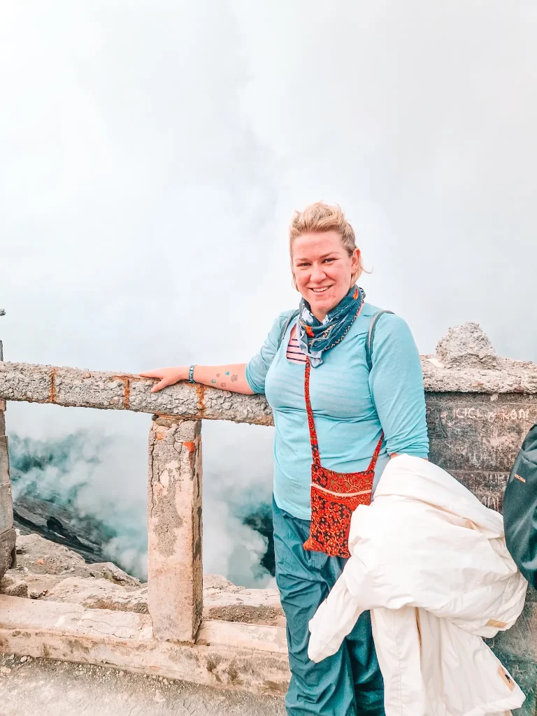 A smiling woman in a blue long-sleeve shirt, navy pants, and a patterned bandana stands near a rocky ledge with a thick cloud of steam rising from below. She leans against a weathered stone railing with her left arm while holding a white jacket in her right hand. A red crossbody bag with intricate patterns hangs from her shoulder.