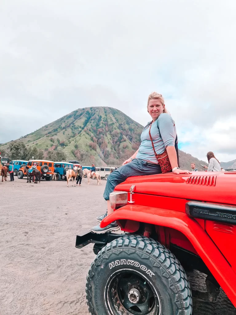 A smiling woman sits atop the hood of a red off-road jeep, posing with one hand on the vehicle. Behind her, a green mountain rises, while colorful jeeps and horses fill the sandy terrain, suggesting a popular adventure destination.