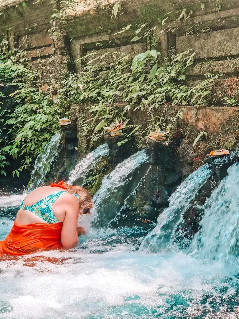 Em wearing a green bikini top and an orange sarong kneels in a sacred Balinese water temple, bowing her head under a stream of water. The stone wall behind her is covered in lush green moss and ferns, with small woven offerings of flowers and leaves placed above each flowing spout. The turquoise water swirls around her as part of a traditional purification ritual.