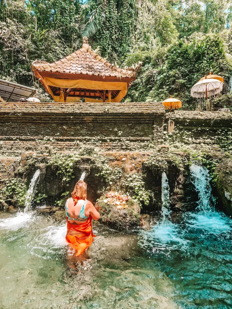 Em wearing a bikini top and an orange sarong wades through clear turquoise water at a sacred Balinese water temple. Small waterfalls flow from an aged stone wall adorned with greenery, and offerings of flowers and leaves rest on a rock. A traditional thatched-roof pavilion and ceremonial umbrellas stand in the background, surrounded by lush tropical foliage.