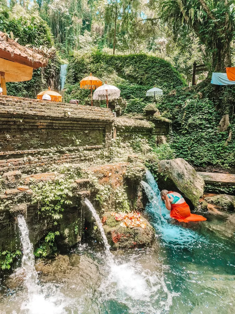 Em dressed in a vibrant orange sarong kneels under a small waterfall, performing a purification ritual at a sacred Balinese water temple. The setting is lush with greenery, moss-covered stone structures, and traditional Balinese umbrellas adorning the area. The cascading water and serene atmosphere highlight the spiritual and natural beauty of this sacred place.